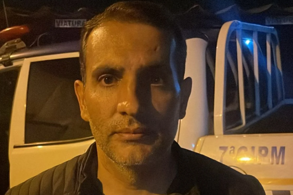 foto de edney a noite, após ser pego pela polícia. ele olha para a câmera, com viatura completando o fundo.